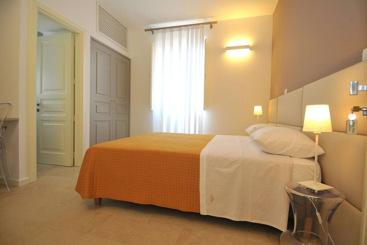 A hotel room with a bed, a bedside table with a lamp, a window with curtains, and a door.