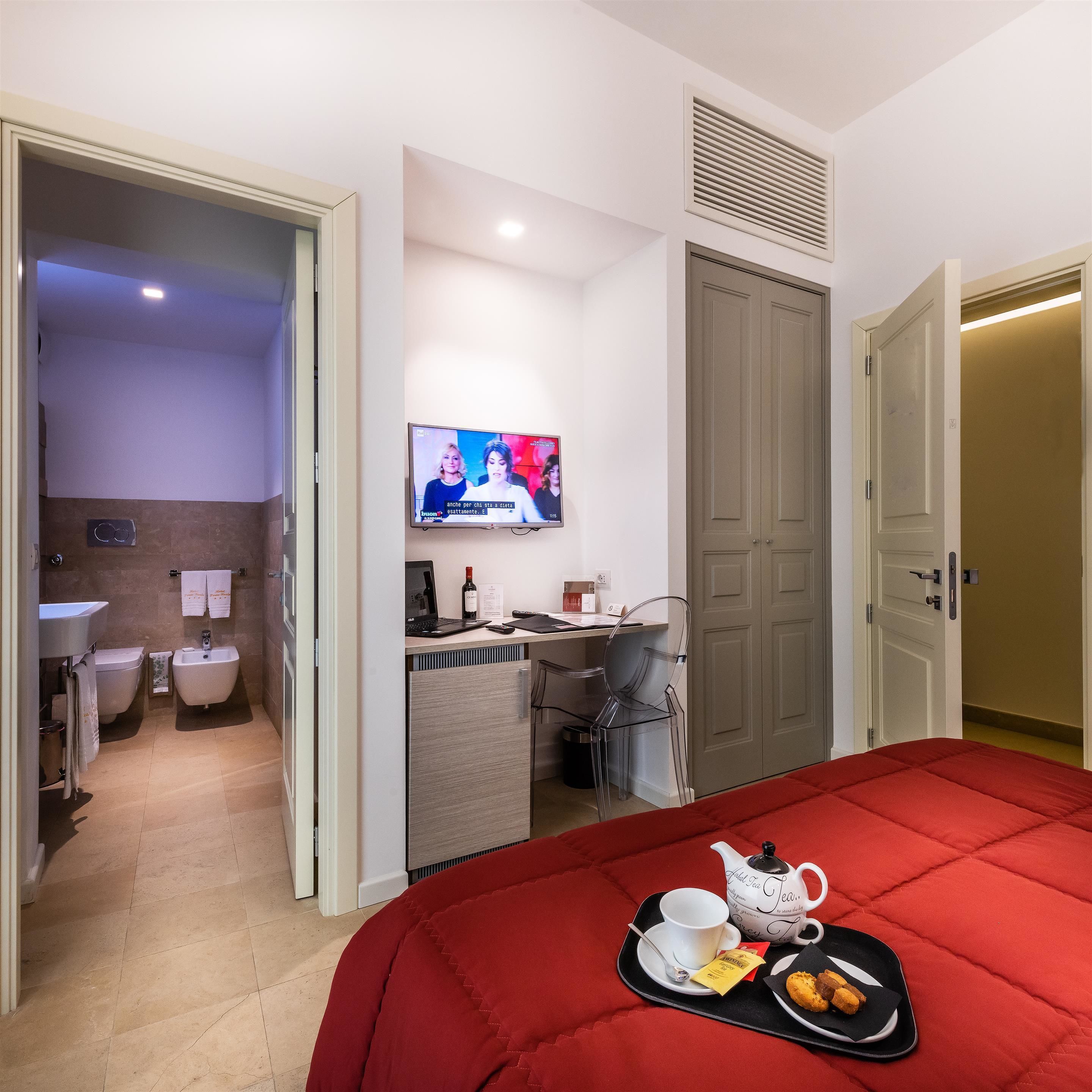 A hotel room with a bed, a desk with a computer, a TV, and a bathroom visible through an open door.