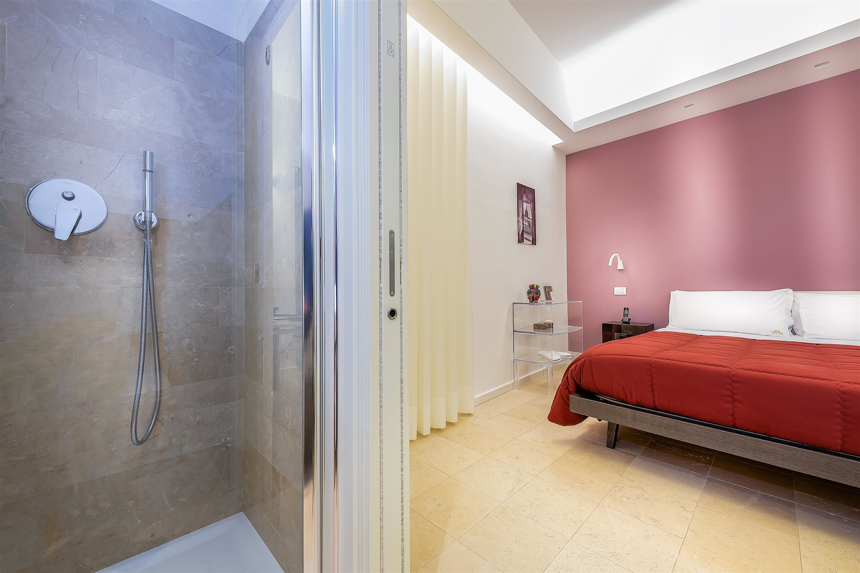 Un bagno moderno con box doccia in vetro, un letto con copripiumino rosso e un comodino con lampada.