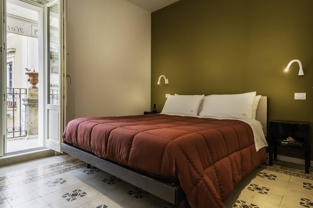A bedroom with a bed, a nightstand, a lamp, and a view of the outside.
