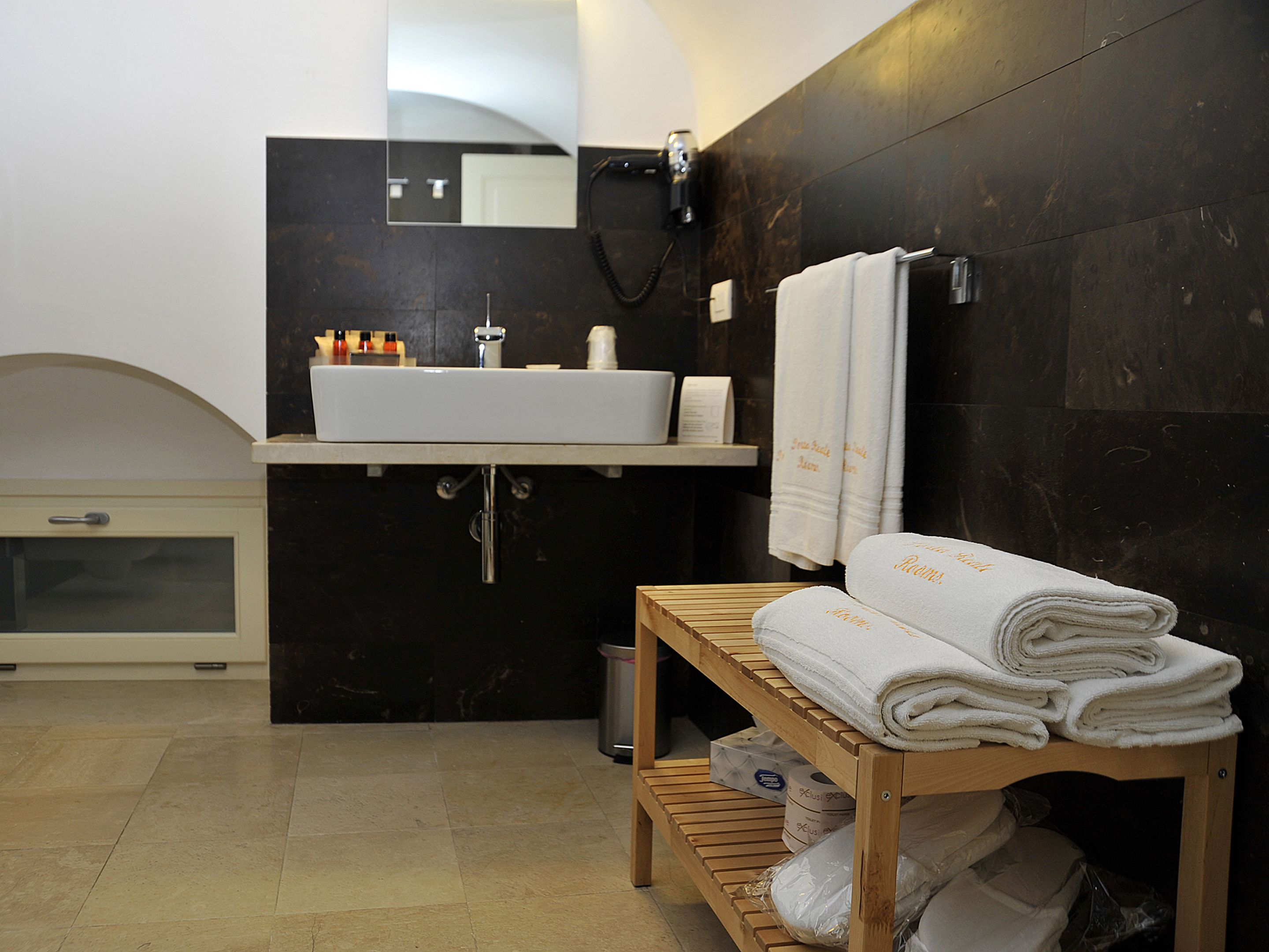 A modern bathroom with a white sink, dark tiled walls, a wooden bench with towels, and a mirror above the sink.