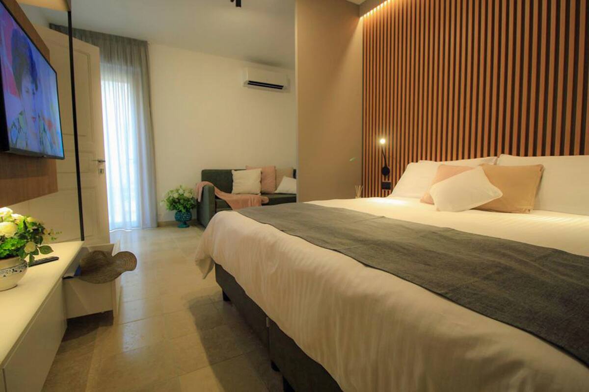 A modern hotel room with a large bed, a flat-screen TV mounted on the wall, a bedside table with a vase of flowers, and a wooden panel wall.