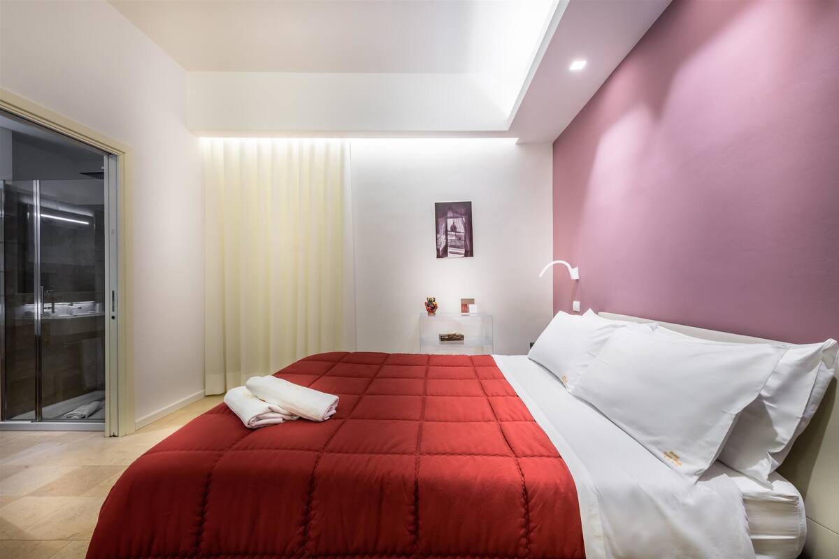 A modern bedroom with a red bedspread, white pillows, a glass shower enclosure, a bedside table with a small lamp, and a framed picture on the wall.