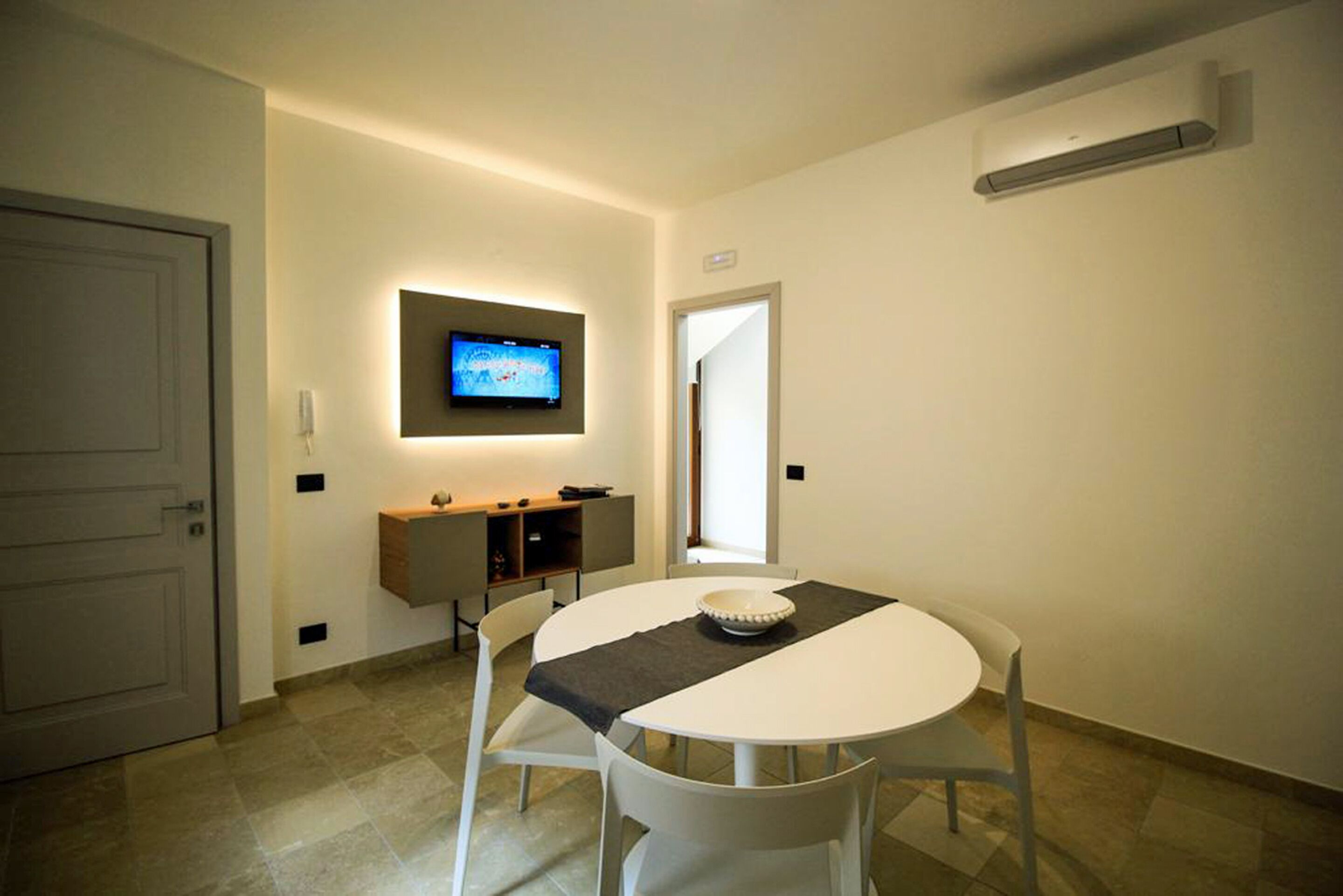 Een moderne eethoek met een ronde tafel, witte stoelen, een flatscreen televisie aan de muur en een kleine kitchenette.