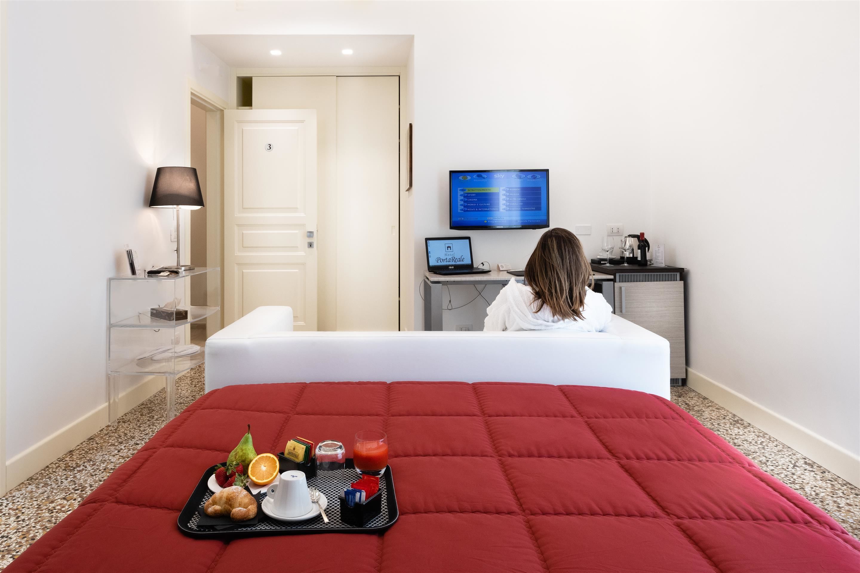 Habitación de hotel con cama, televisión, escritorio con una computadora portátil y una bandeja con alimentos para el desayuno.
