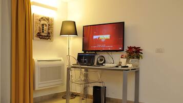 Ett litet, väl upplyst rum med ett skrivbord, en stol, en tv som visar SKY MUSIC, en lampa, ett element och en inramad tavla på väggen.