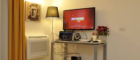 Una stanza piccola e ben illuminata, con una scrivania, una sedia, un televisore che mostra SKY MUSIC, una lampada, un termosifone e un quadro appeso al muro.