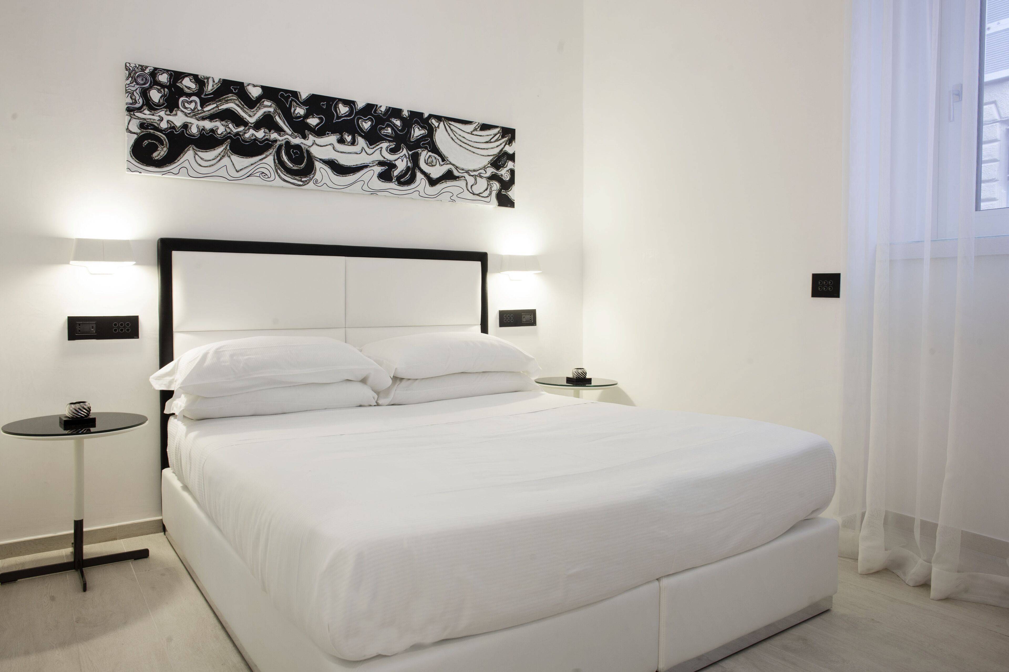 Ein modernes Hotelzimmer mit einem großen Bett, einem Kopfteil mit integrierter Beleuchtung, einem kleinen Nachttisch und einem abstraktem Kunstwerk an der Wand.