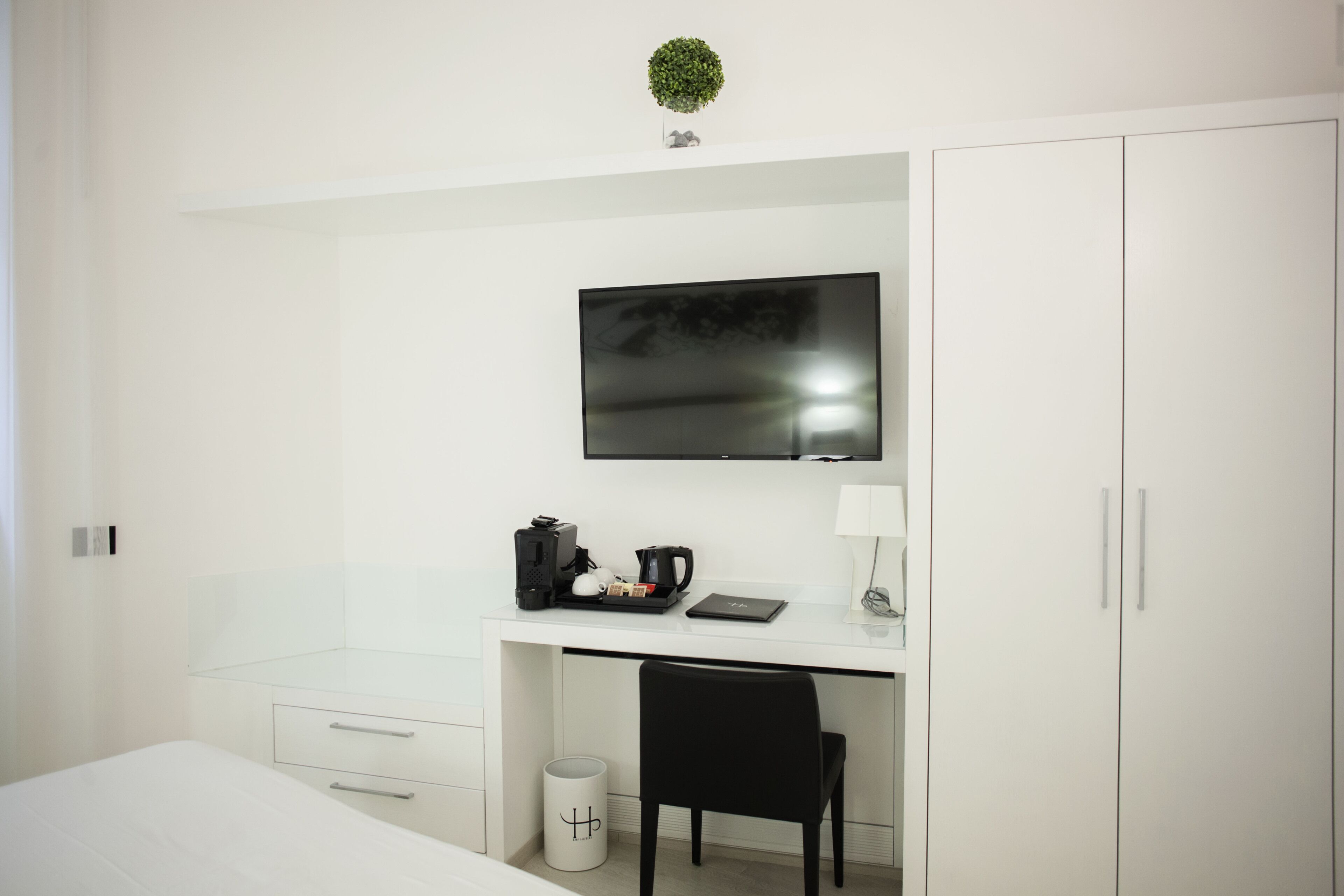 Habitación de hotel moderna con un escritorio blanco, un televisor de pantalla plana, una silla negra y una pequeña planta en maceta en la pared.