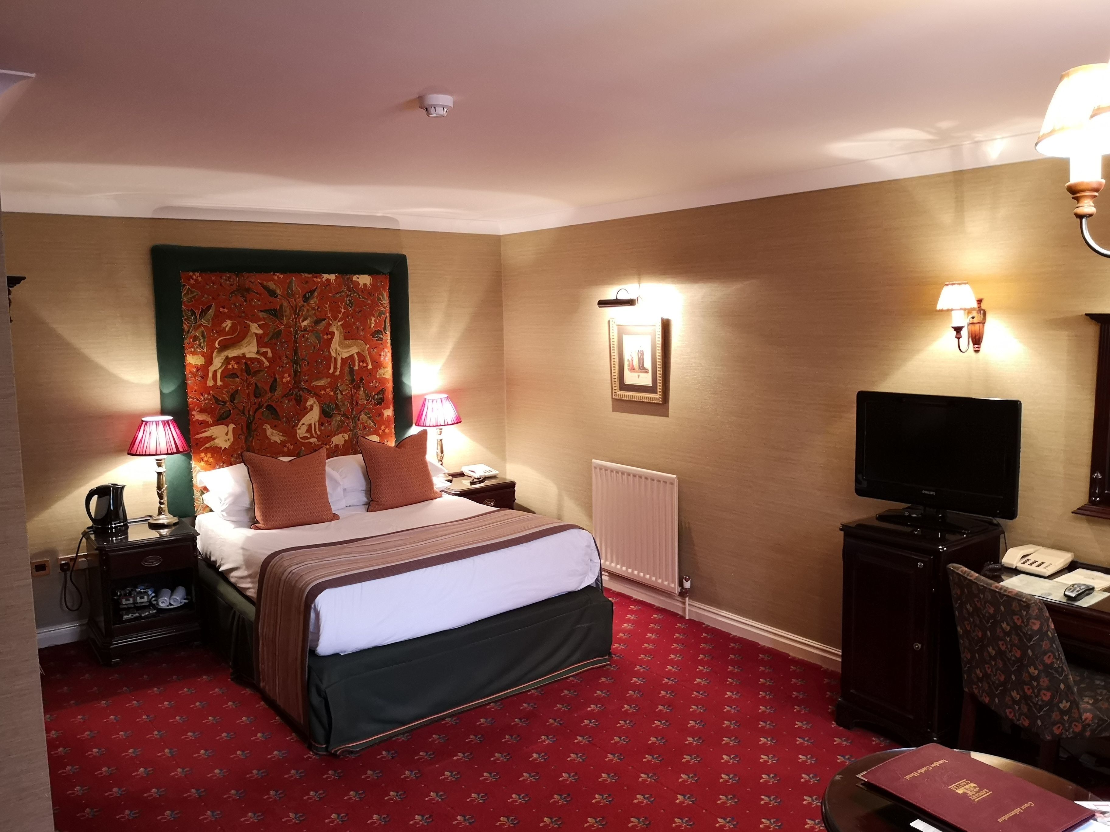 A hotel room with a large bed, two bedside lamps, a television, a desk with a telephone, and a radiator.