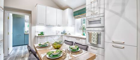 Una cocina moderna con armarios blancos, una mesa de comedor de madera dispuesta para cuatro comensales y un horno empotrado.