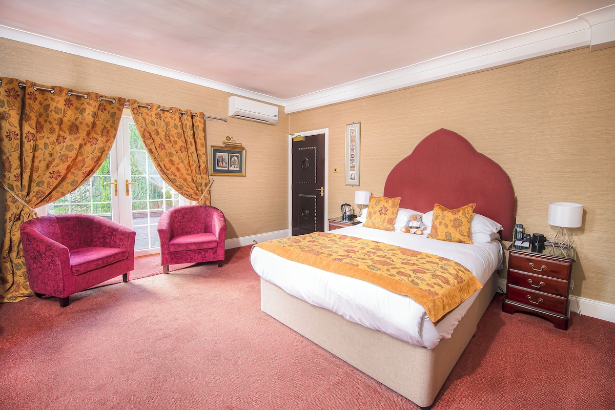 A hotel room with a large bed, two red armchairs, a wooden nightstand, and a window with curtains.