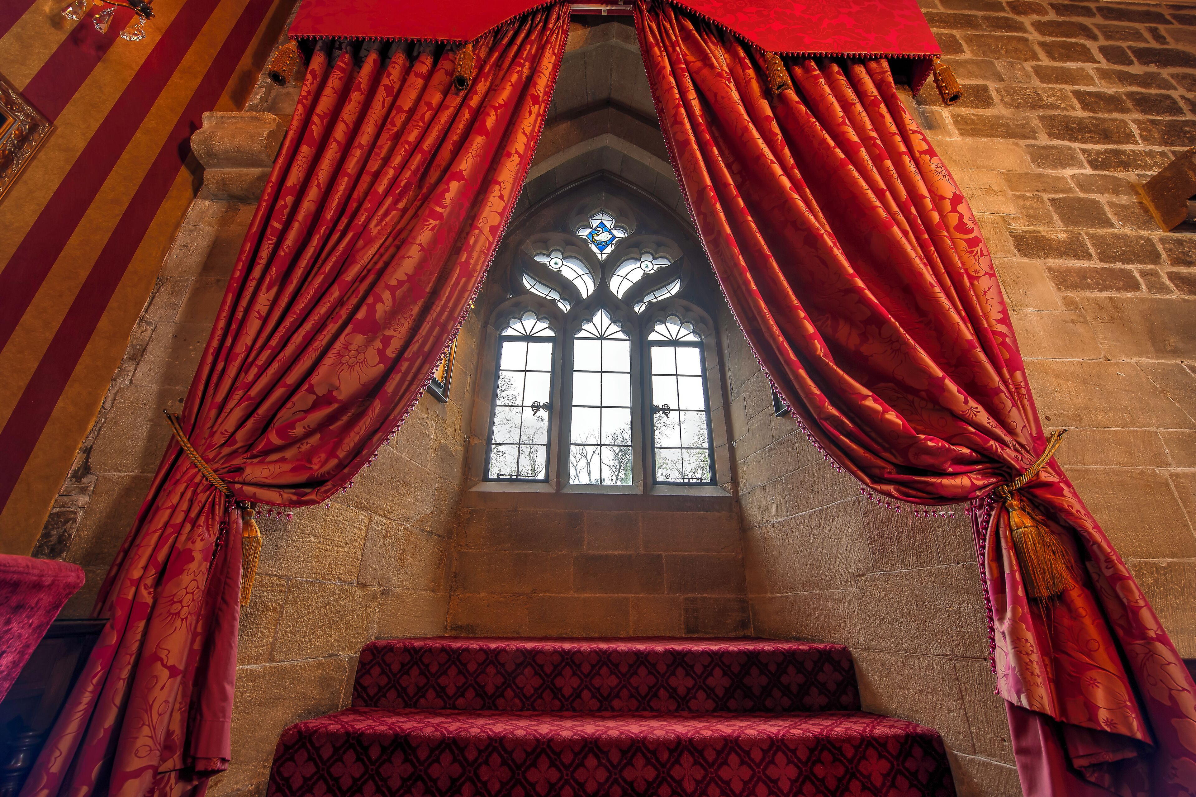 Una ventana de estilo gótico con cortinas rojas, paredes de piedra y una zona alfombrada con diseño.
