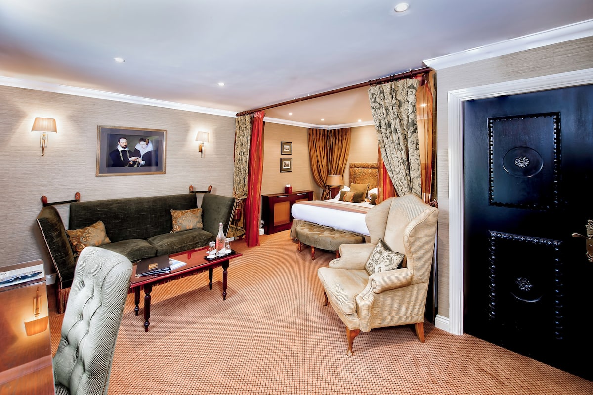 A hotel room with a sofa, armchair, and a bed. There is a picture on the wall and a lamp on the side.