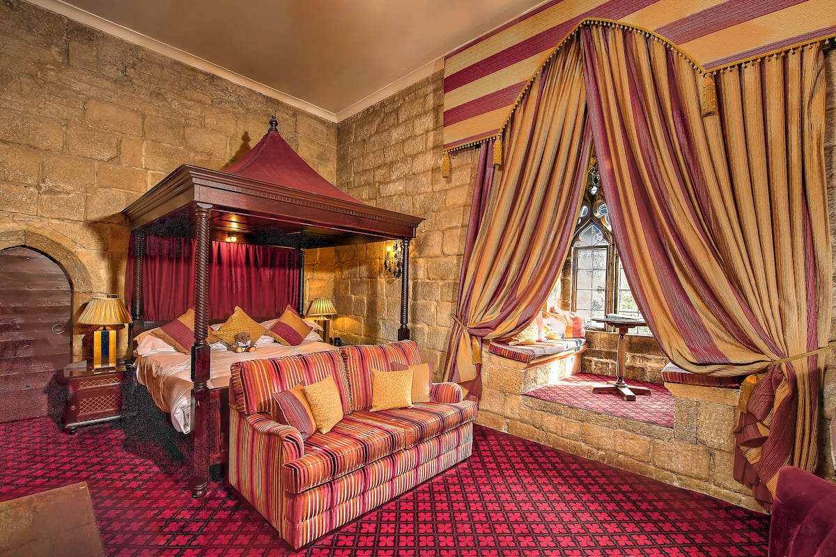 A room with a four-poster bed, a sofa, a stone wall, and a window with curtains.