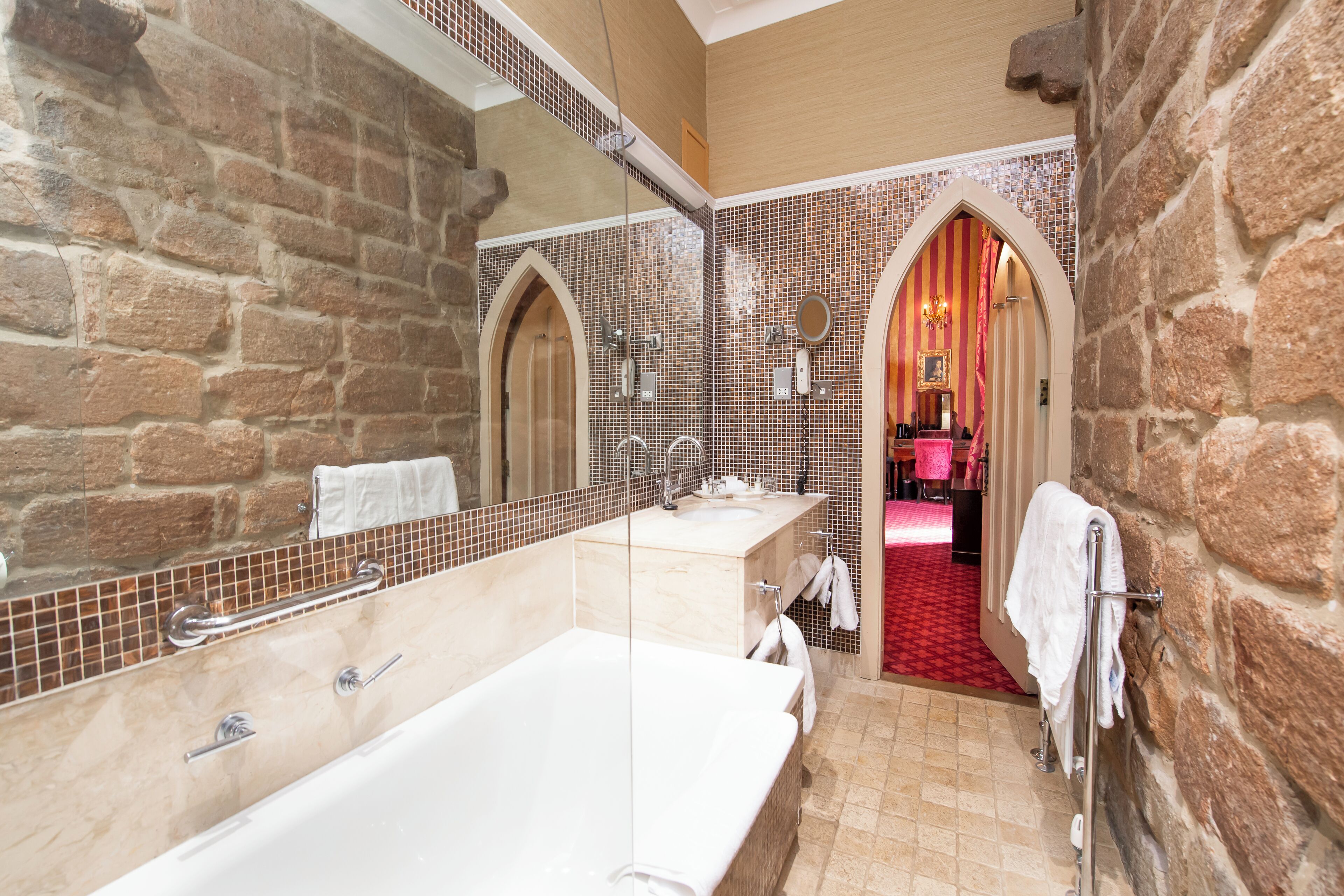 Un baño con una bañera grande, un lavamanos y un espejo. Hay un perchero y una alfombra roja que conduce a una habitación con pared rosa.