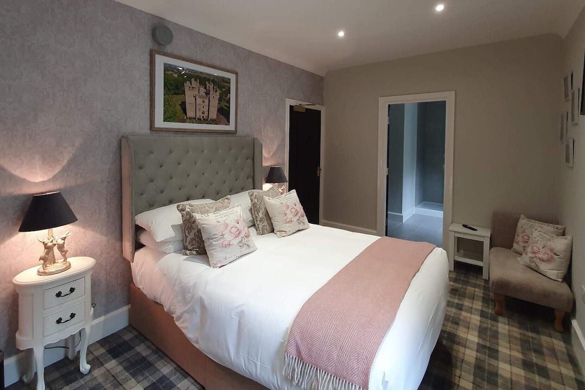 A bedroom with a large bed, a nightstand with a lamp, a chair, and a door leading to another room.