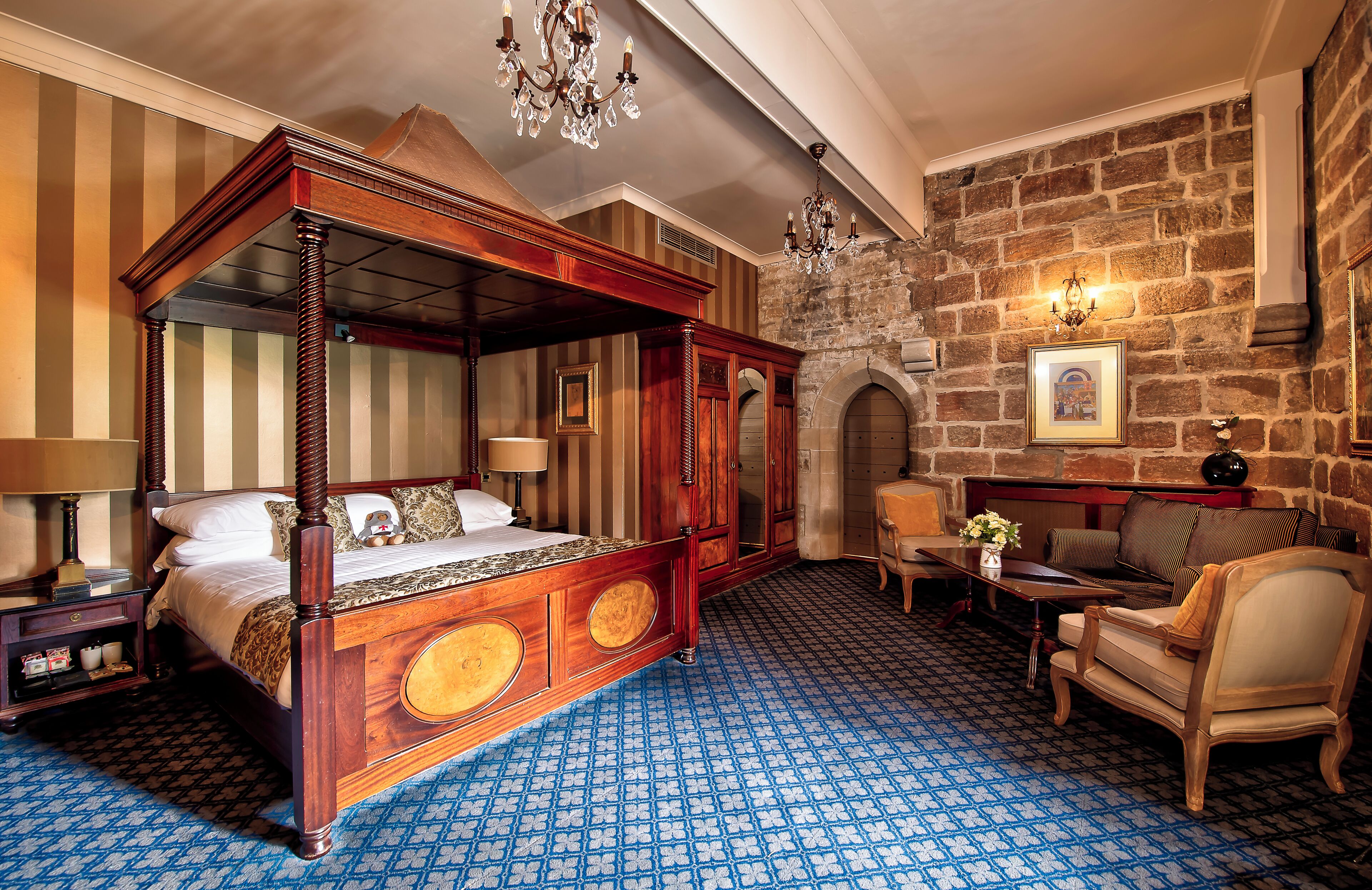 A four-poster bed with a wooden frame and a blue patterned carpet. There is a stone wall with a fireplace and a chandelier.