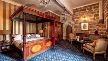 Una cama con dosel de madera y una alfombra azul estampada. Se observa una pared de piedra con chimenea y un candelabro.