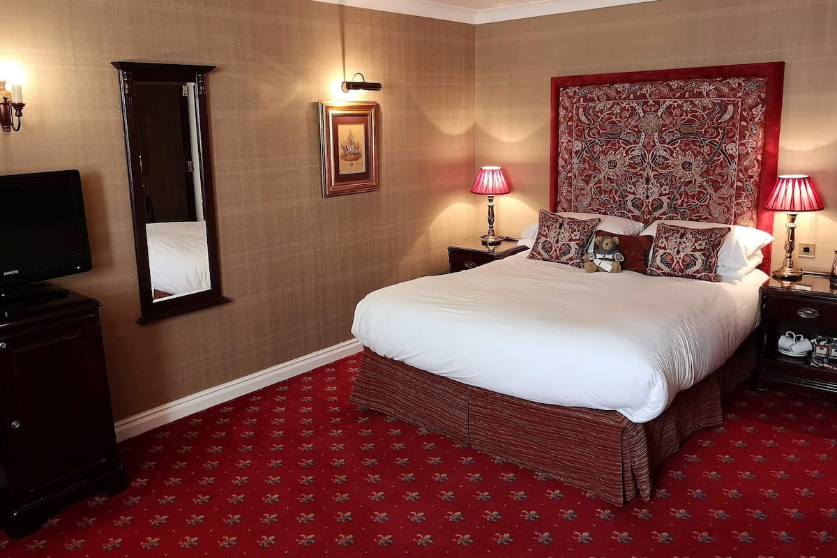 A hotel room with a large bed, two bedside tables with lamps, a flat-screen TV, a large mirror, and a patterned wall hanging.
