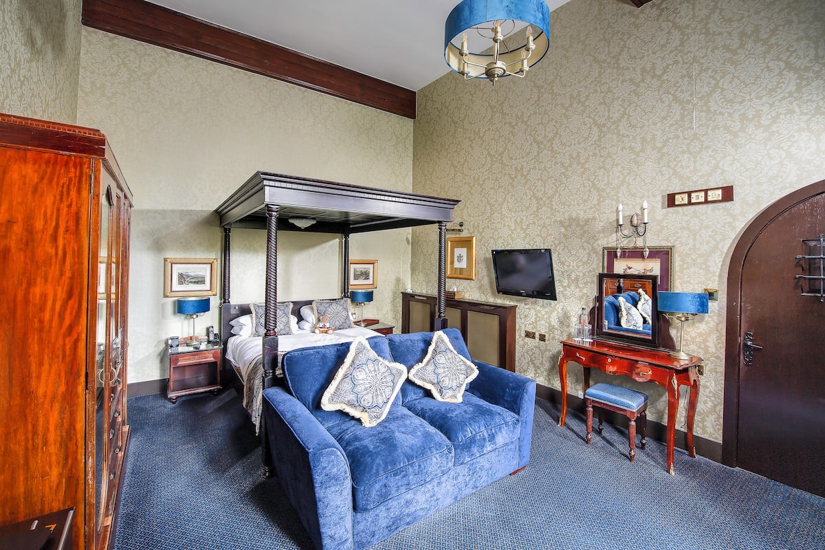 A bedroom with a four-poster bed, a blue sofa, a wooden desk, and a television.
