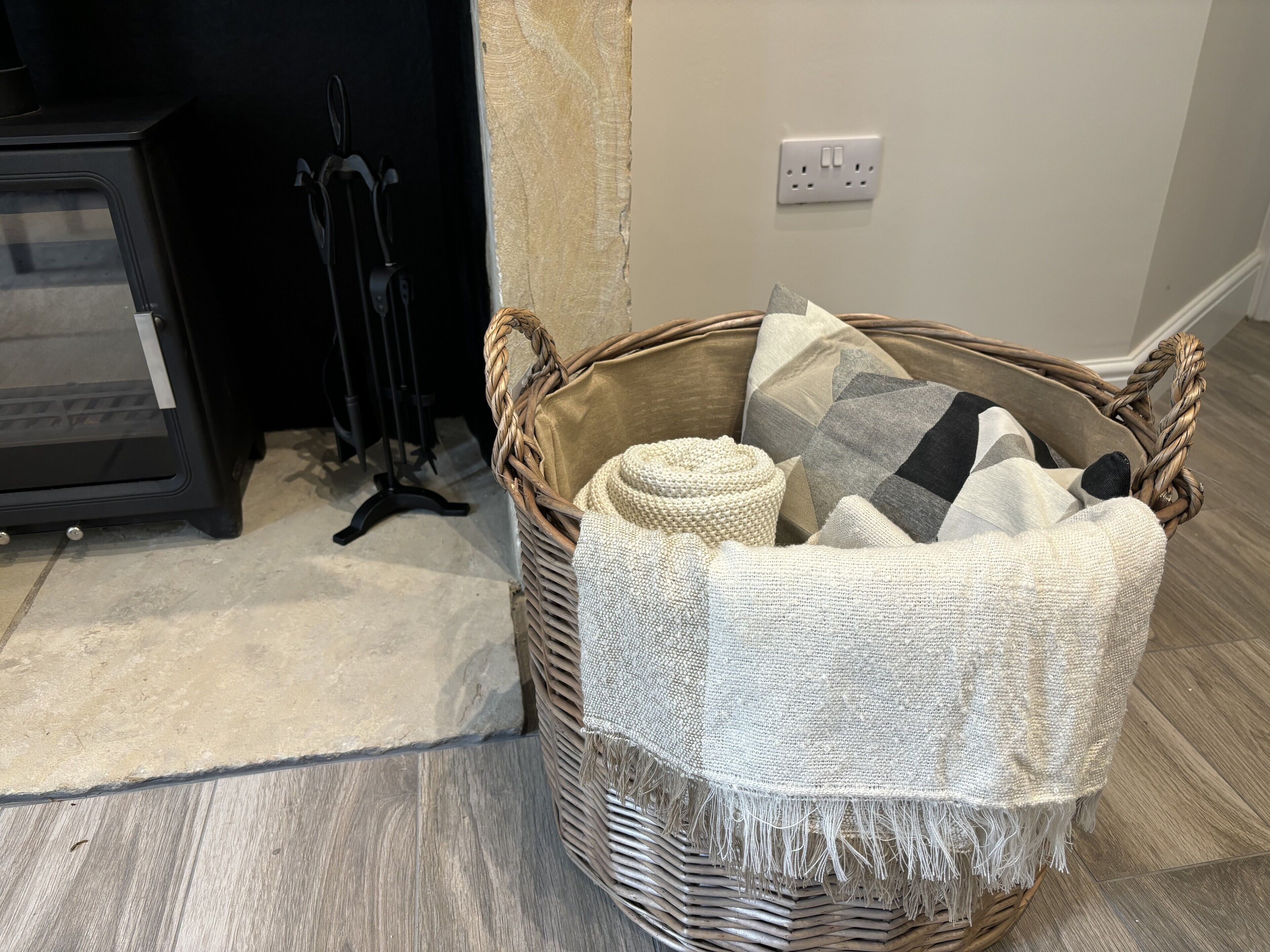 A wicker basket with a woven blanket and pillows on a wooden floor.