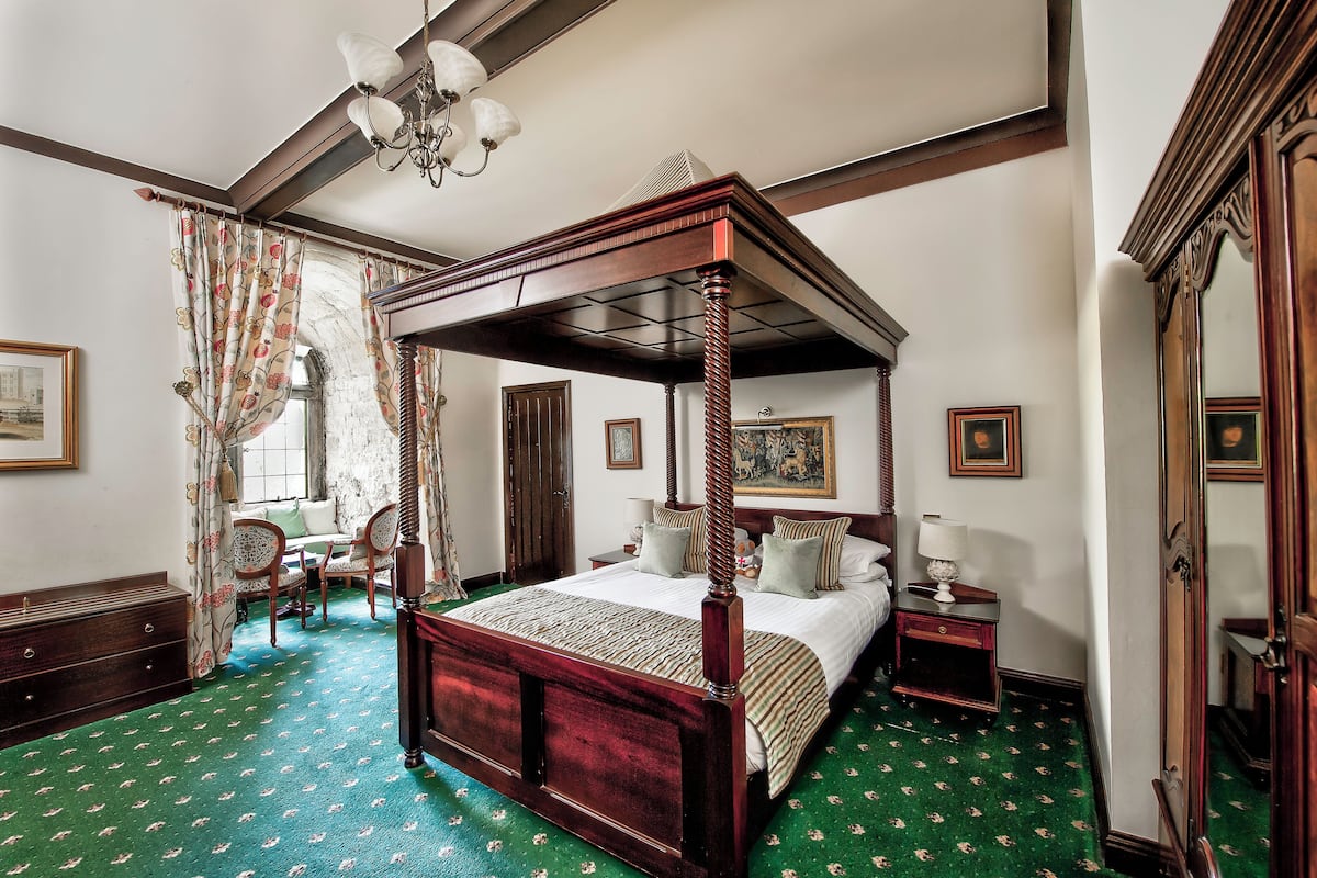 A four-poster bed with a wooden frame and a green carpeted floor. There is a nightstand with a lamp and a chair with a cushion.