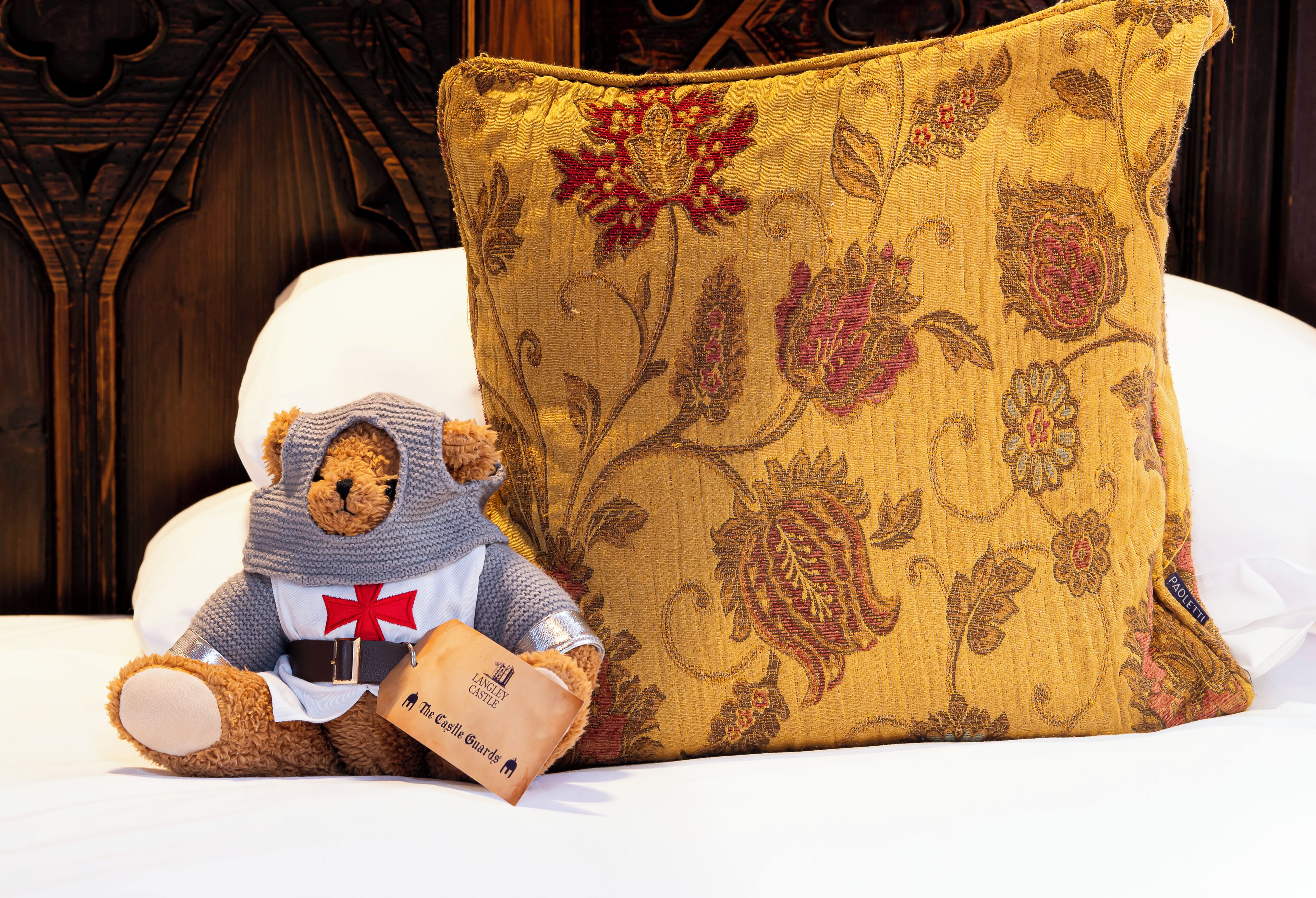 A teddy bear in a knitted outfit sitting on a bed with a floral-patterned pillow.