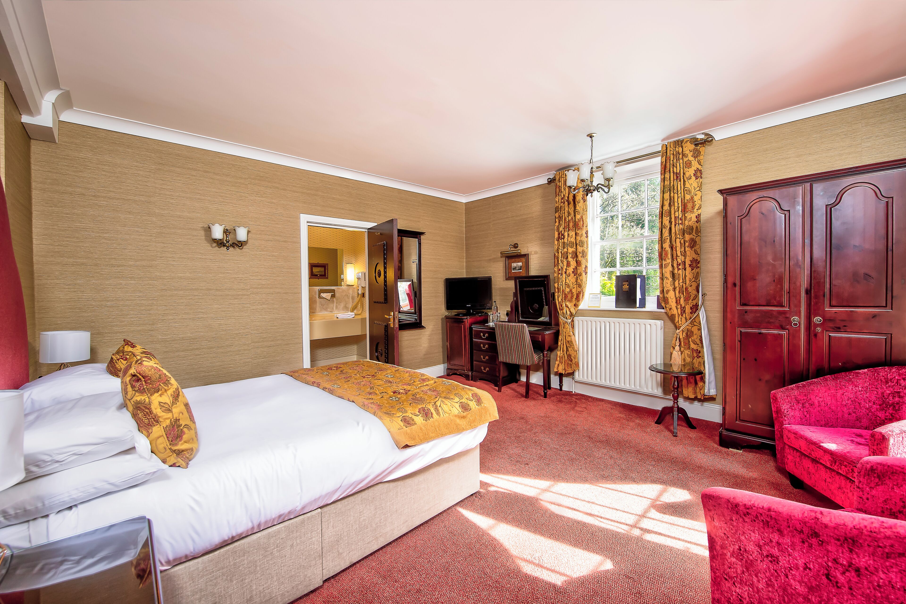 A hotel room with a bed, a desk, a chair, a red armchair, and a wardrobe.