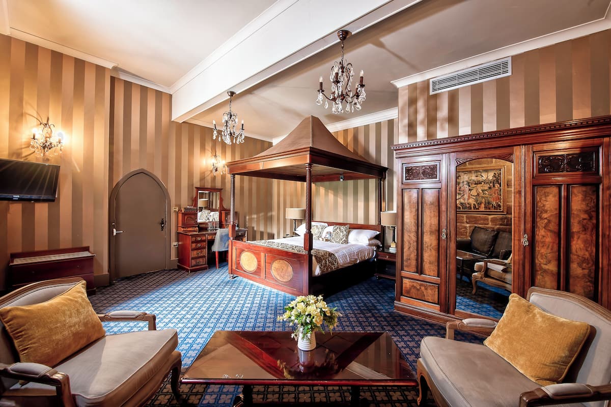 A hotel room with a four-poster bed, a sitting area with two armchairs, a wooden cabinet, and a television on the left.