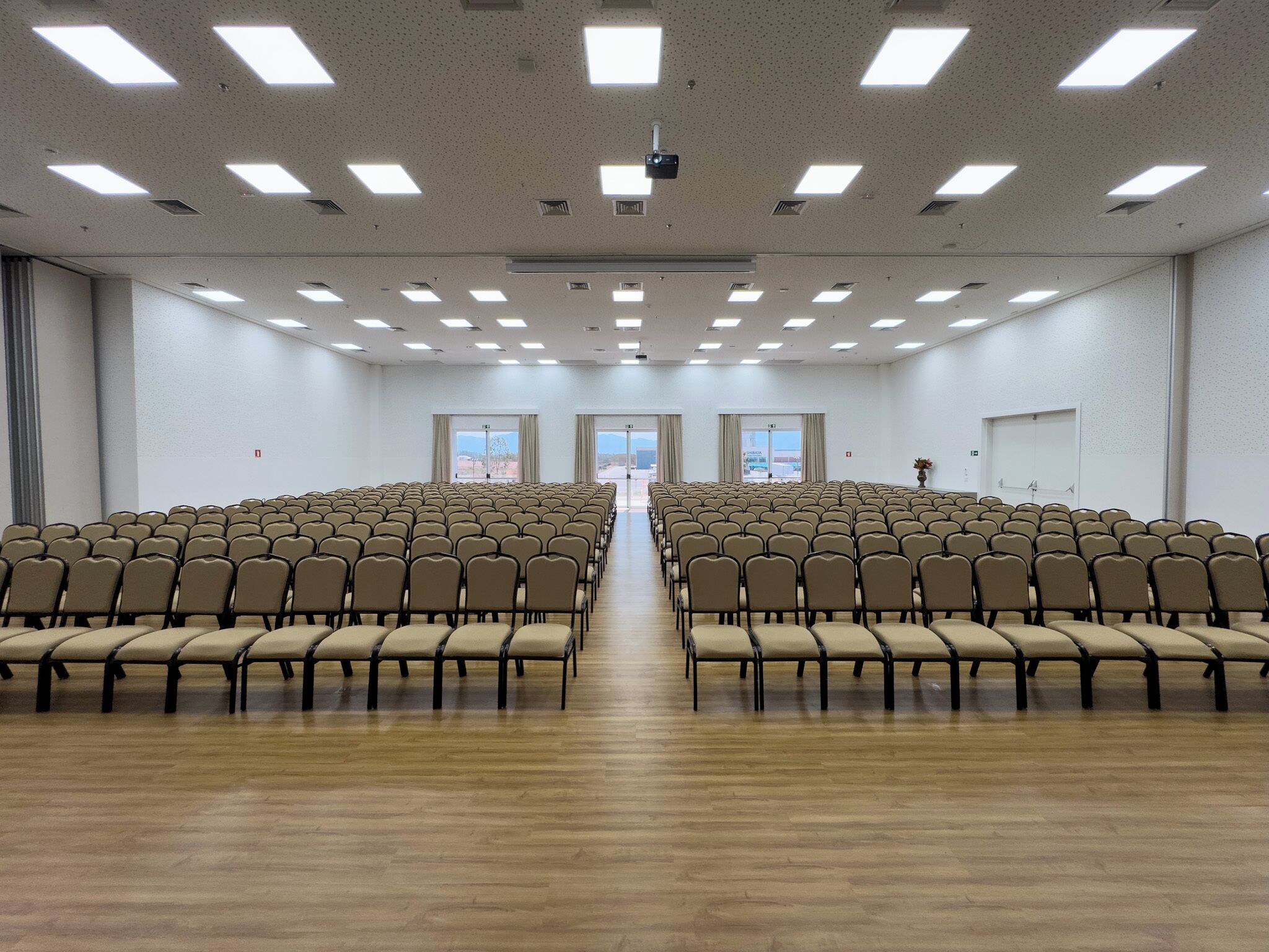 Un salón de conferencias con filas de sillas, un proyector y ventanas amplias.