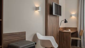 Habitación de hotel moderna con cabecera de madera, televisor de pantalla plana montado en la pared, escritorio con silla, maleta y lámpara.