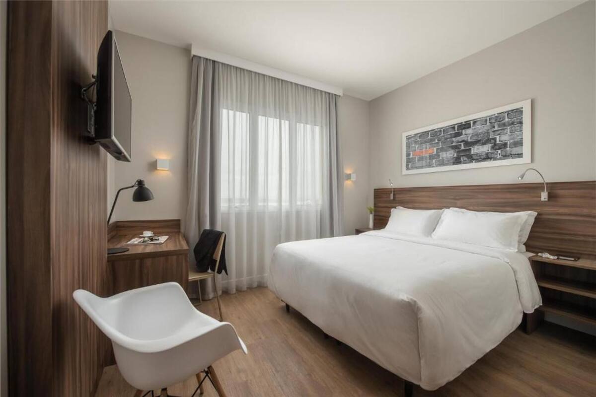 A hotel room with a bed, a desk, a chair, a TV, and a window with curtains.