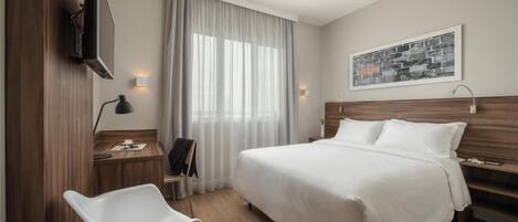 Habitación de hotel con cama, escritorio, silla, televisor y ventana con cortinas.