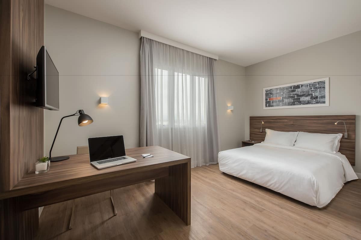 A modern hotel room with a wooden desk, a laptop, a bedside lamp, a bed with white bedding, a mounted TV, and a framed picture on the wall.