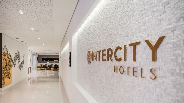 Un pasillo con piso pulido que conduce a una recepción con pared texturizada y el texto 'INTERCITY HOTELS'.