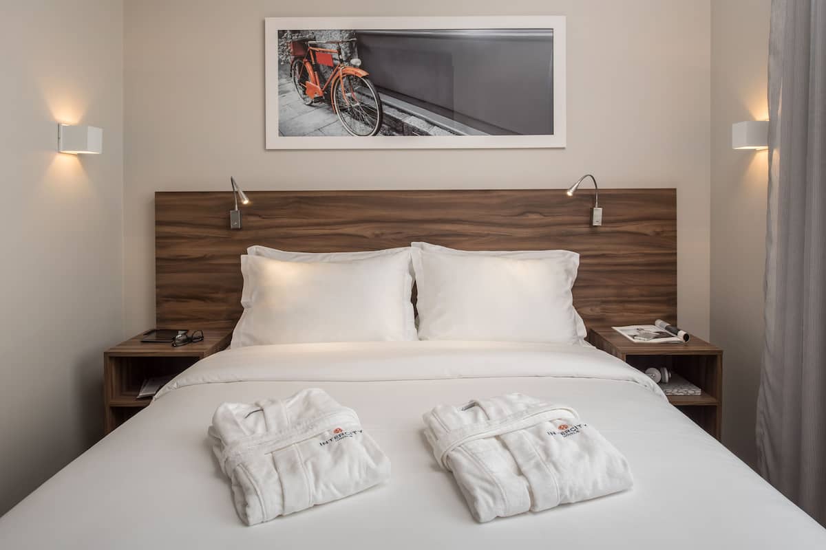 A neatly made bed with two white bathrobes, a wooden headboard, bedside tables with lamps, and a framed picture above the bed.