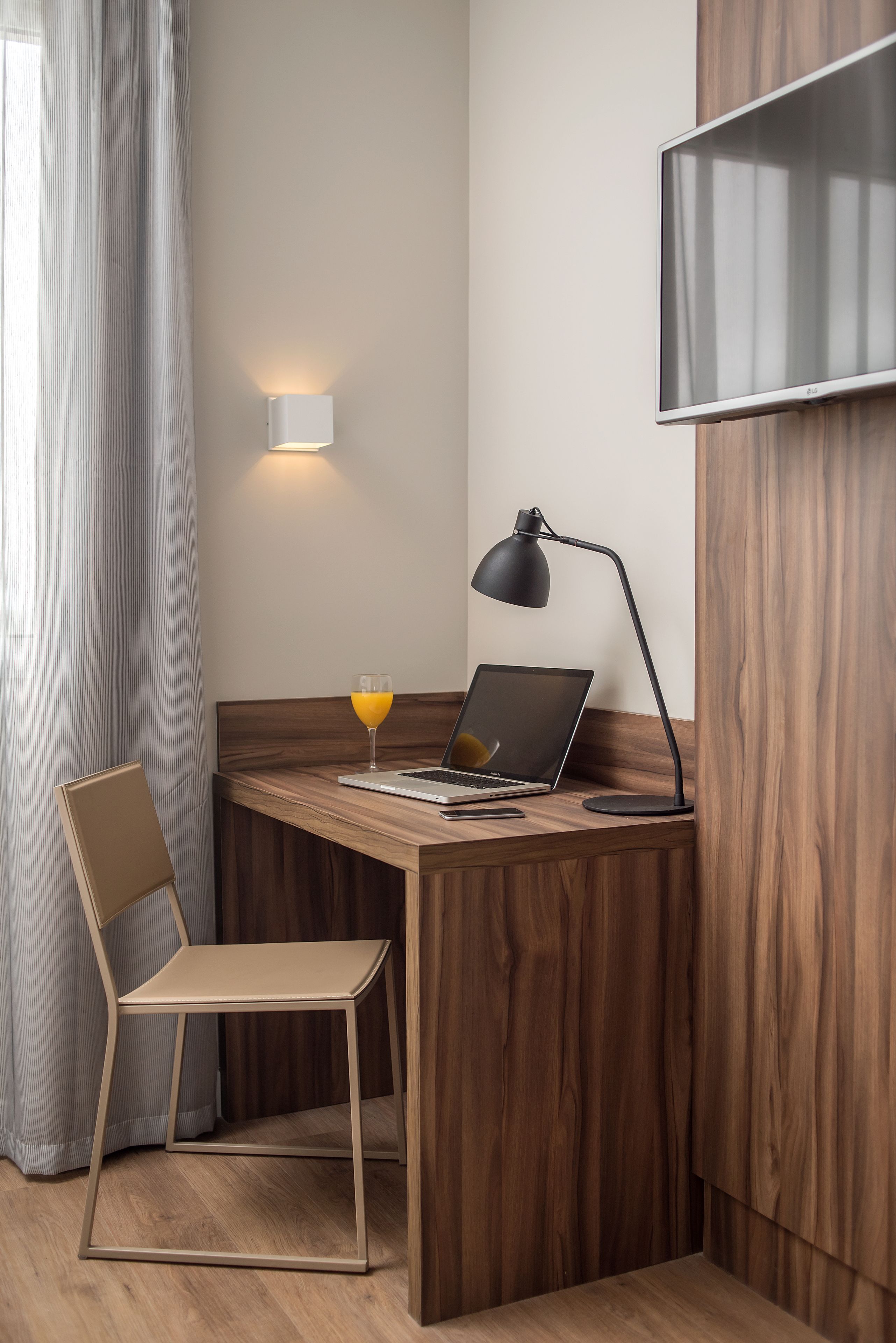 A modern workspace with a wooden desk, a laptop, a glass of orange juice, a chair, and a wall-mounted TV.