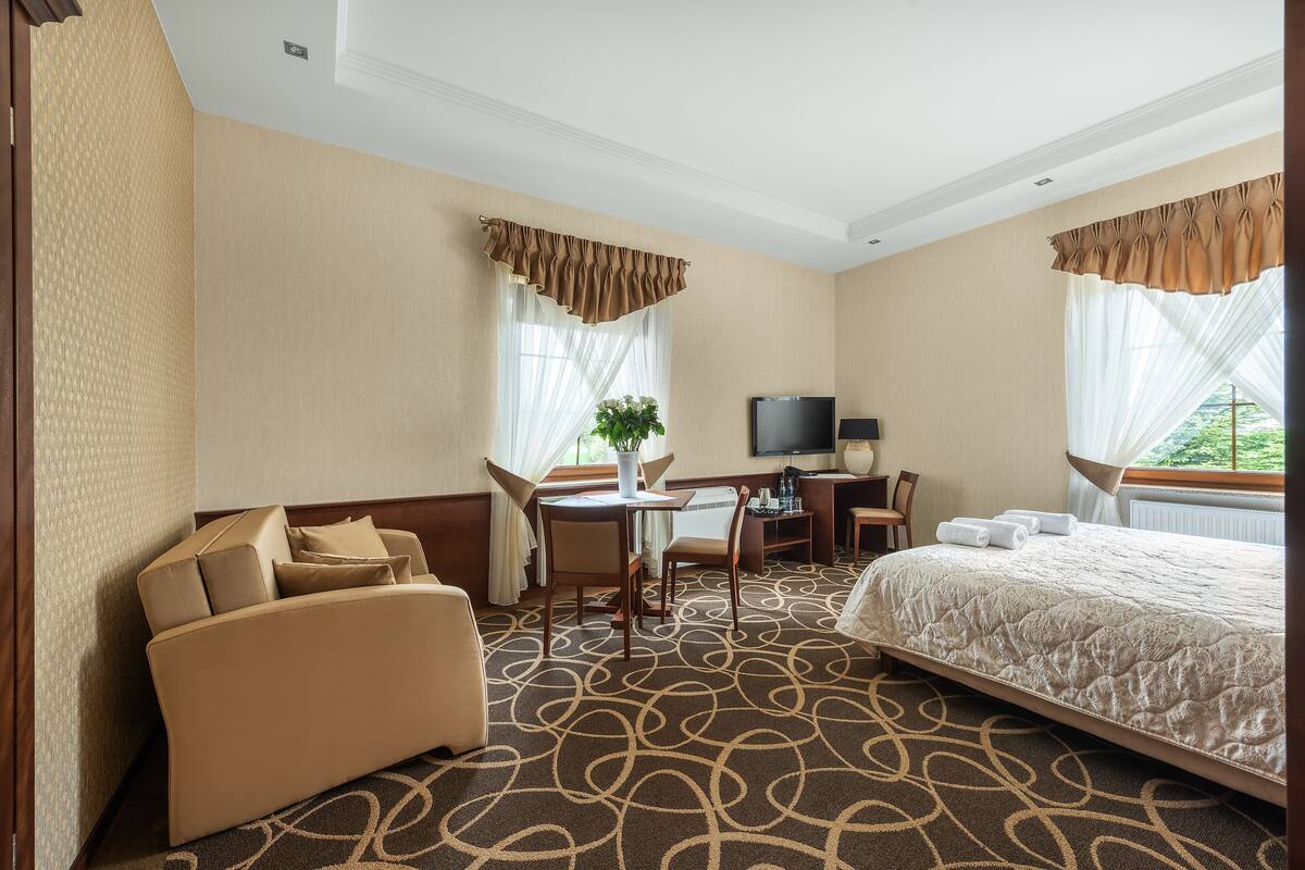 A hotel room with a bed, a sofa, a desk with a chair, a television, and a window with curtains.