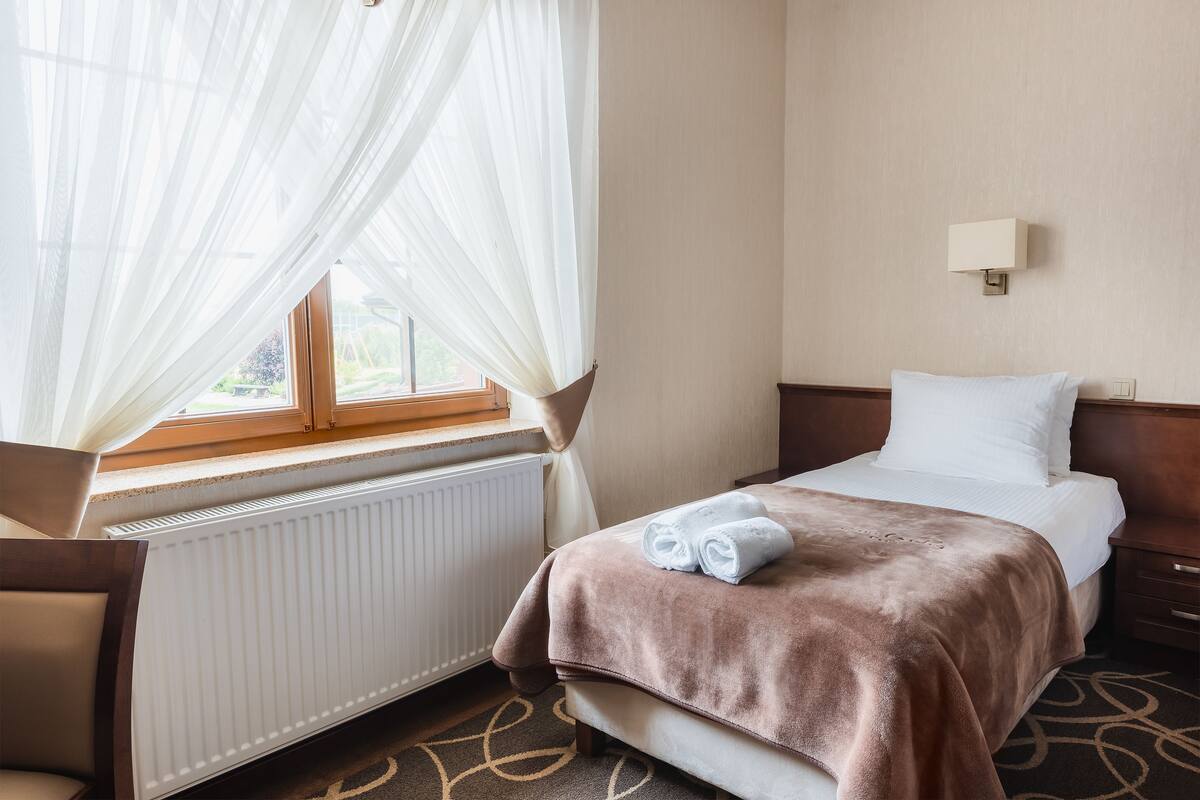 A hotel room with a bed, a bedside table, a lamp, a window with sheer curtains, and a radiator.