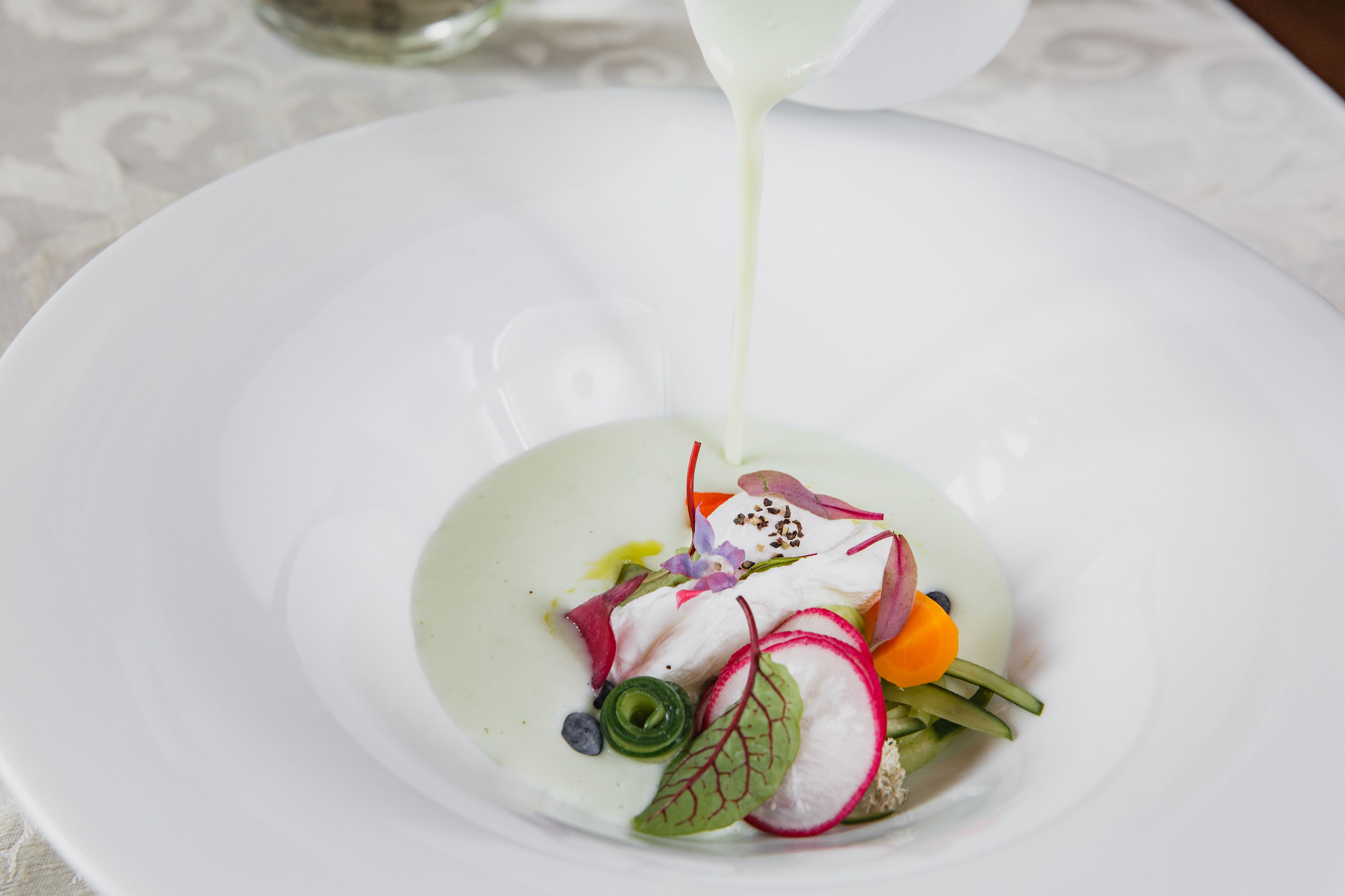 A white plate with a white sauce being poured over a dish of fish and vegetables.