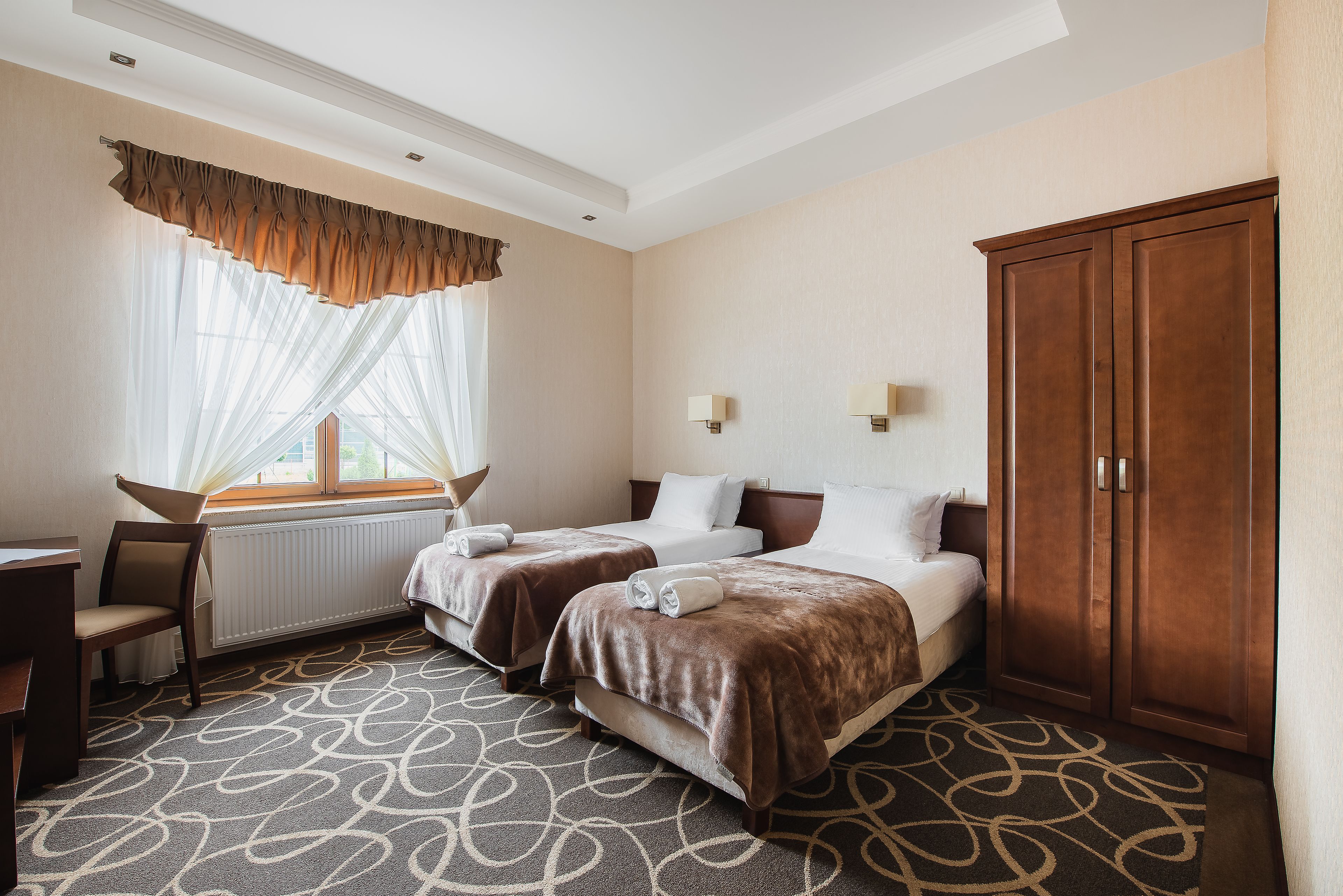 Une chambre d’hôtel avec deux lits, un bureau, une chaise, une armoire et une fenêtre avec des rideaux légers.