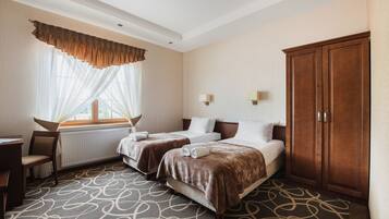 Une chambre d’hôtel avec deux lits, un bureau, une chaise, une armoire et une fenêtre avec des rideaux légers.