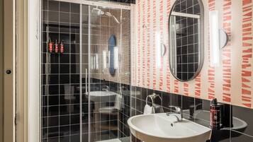 Un baño moderno con cabina de ducha de vidrio, un lavabo blanco y un espejo frente a una pared con estampado rojo y blanco.