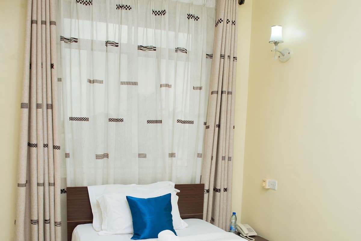 A neatly made bed with a blue and white bedspread, a wooden headboard, and a white wall with a light switch and a small table with a phone and a bottle.