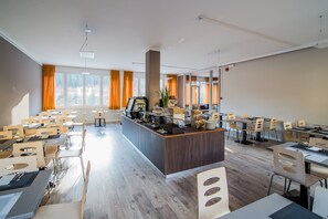 A modern dining area with multiple tables and chairs, a central counter with a coffee machine, and large windows with orange curtains. - Hotel alla fonte (Arta Terme)