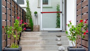 Une série de marches en béton menant à une porte d’entrée en verre, flanquée de clôtures en bois et de plantes en pot ornées de fleurs rouges.