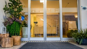 L’entrée d’un hôtel, avec une porte en verre, des plantes en pot et un banc en bois.