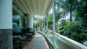 A covered outdoor seating area with tables and chairs, surrounded by glass railings and tropical plants. - Ubumwe Grande Hotel (Kigali)