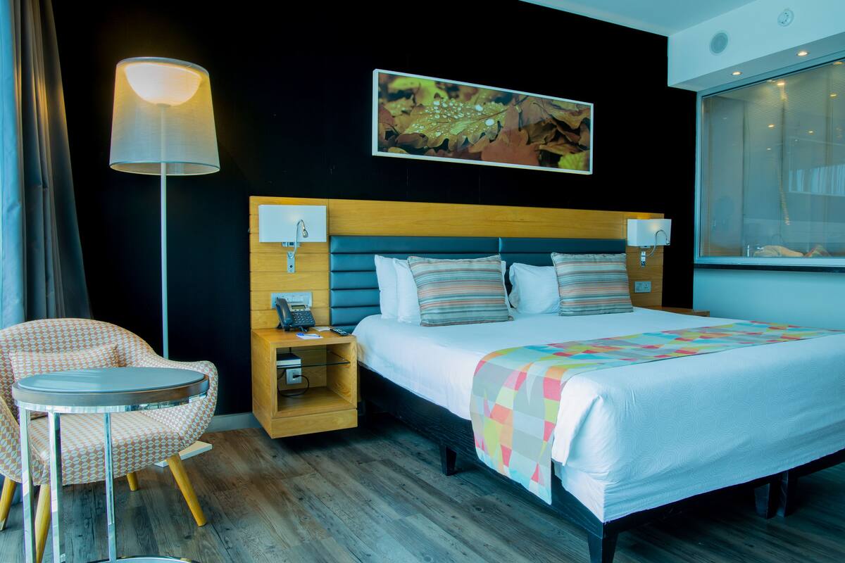 A hotel room with a bed, bedside table, lamp, chair, and artwork on the wall.